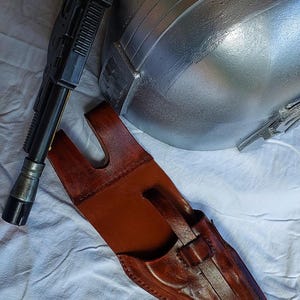 May include: A brown leather holster with a black blaster and a silver helmet. The holster is designed to hold the blaster. The helmet is metallic and has a smooth surface. The blaster is black and has a detailed design.