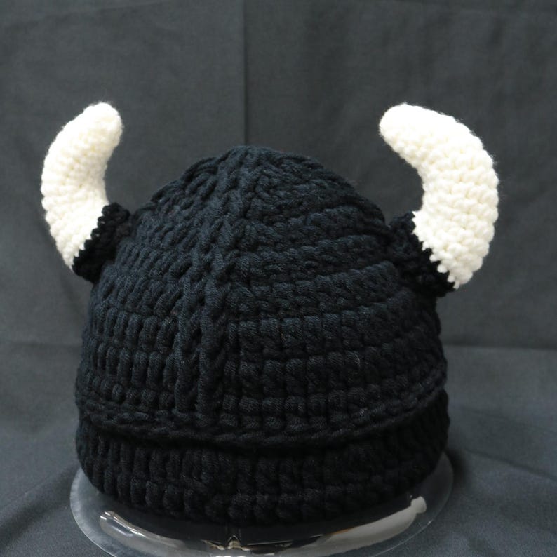 May include: A black crocheted beanie hat featuring two prominent, curved white horns attached to the sides. The hat is constructed with a thick, textured yarn pattern and has a solid black base with a ribbed edge, mimicking a horned helmet design.