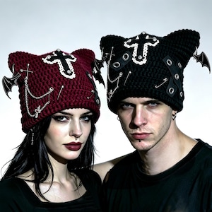 May include: Two crochet cat-ear beanies, one in deep red and one in black. Each features a white-trimmed cross patch, silver safety pins, dangling chains, metal eyelets with crisscrossed straps, and small black bat-shaped charms on the sides.