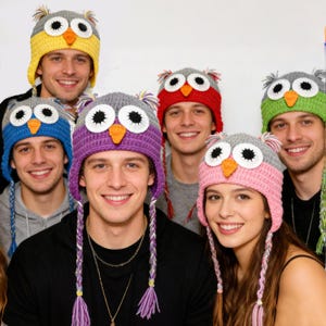 May include: A group of people wearing colorful, handmade owl-themed crochet hats. The hats feature large eyes, orange beaks, and ear flaps. The hats are in various colors, including yellow, blue, red, purple, green, and pink.