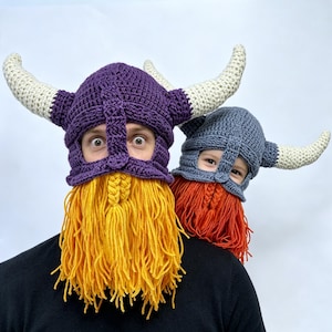 May include: Two crocheted Viking hats with horns and attached yarn beards. The larger hat is purple with a bright yellow beard, while the smaller hat is gray with a vibrant orange beard. Both feature cream-colored horns and face openings.
