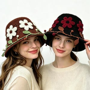 Puede incluir: Dos sombreros de pescador de ganchillo, uno marrón con flores blancas y enredaderas verdes, y otro negro con flores rojo oscuro y enredaderas marrones. Ambos sombreros presentan aplicaciones detalladas de flores y hojas cosidas.