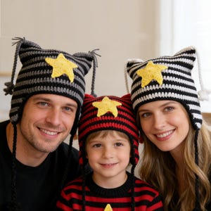 May include: Three people wearing matching crochet hats with star accents. The hats feature ear-like flaps and striped patterns in black and grey, black and white, and red and black. A yellow star is centered on each hat.