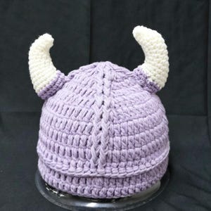 May include: A light purple crocheted hat featuring two prominent, curved white horns extending from the sides. The hat is constructed with a textured yarn pattern and a vertical braided detail running down the center of the front section.