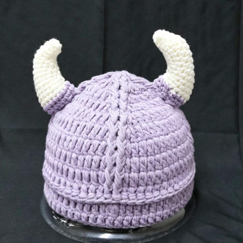 May include: A light purple crocheted hat featuring two prominent, curved white horns extending from the sides. The hat is constructed with a textured yarn pattern and a vertical braided detail running down the center of the front section.