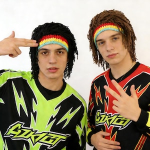 May include: Two young people wearing black dreadlock wigs with colourful headbands. They are wearing black and neon green or red and black jerseys with a lightning bolt design and the word "SKGT". One person is pointing their fingers like a gun.