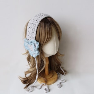May include: A crocheted gray headband with a light blue flower accent. The headband has a textured pattern and is designed to cover the ears. The flower is light blue with a white center. The headband has two dangling cords.