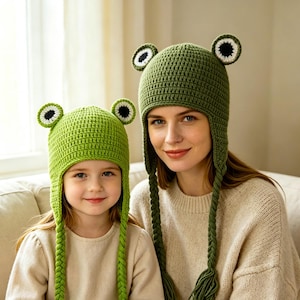 May include: Two matching green crochet hats featuring frog-like eyes on top, ear flaps, and long braided tassels. The hats are constructed with a textured stitch pattern, incorporating white and black circular details to mimic frog eyes.