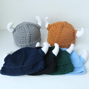 May include: A collection of six crocheted hats, each featuring two white horns. The hats are available in various solid colors including gray, tan, navy blue, black, dark green, and light blue, all crafted with a textured, ribbed yarn pattern.