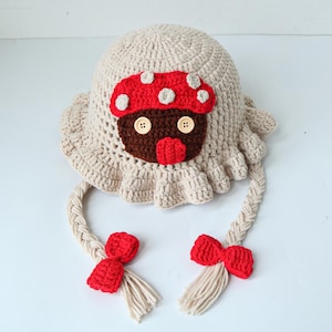 May include: A beige crochet bucket hat featuring a central brown mushroom applique with a red cap, white spots, and two light-colored buttons for eyes. The hat has a ruffled brim and two long braided beige tassels, each adorned with a red crochet bow.