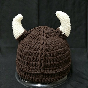 May include: A dark brown crocheted hat featuring a textured vertical cable knit pattern and two prominent, curved white horns extending from the sides. The hat is constructed from thick yarn with a dense, uniform stitch throughout the design.