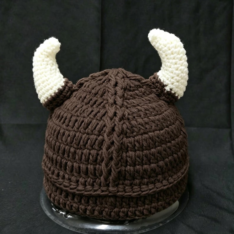 May include: A dark brown crocheted hat featuring a textured vertical cable knit pattern and two prominent, curved white horns extending from the sides. The hat is constructed from thick yarn with a dense, uniform stitch throughout the design.