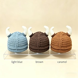 May include: Three crocheted hats with horns, arranged in a row. The hats are light blue, brown, and caramel, each featuring two white curved horns. Below each hat, the text labels light blue, brown, and caramel are clearly visible.