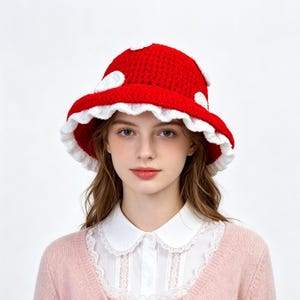 Puede incluir: Un sombrero de pescador de ganchillo rojo brillante con un borde blanco con volantes y parches circulares blancos dispersos por la copa. El sombrero está construido con una textura de hilo visible y tiene una forma redondeada.