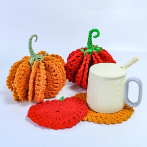 Puede incluir: Dos calabazas de ganchillo en naranja y rojo con tallos verdes, un posavasos de ganchillo rojo, un posavasos de ganchillo naranja y una taza color crema con un patrón de veta de madera clara y una cuchara pequeña.