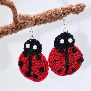 May include: A pair of ladybird earrings crafted from red and black yarn. Each earring features a round red body with black spots, a black head section, and two small white pearl-like beads for eyes. The earrings hang from silver-toned metal hooks.