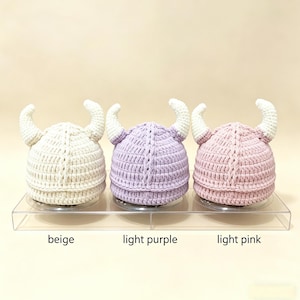 May include: Three crocheted hats with horns, arranged in a row. The hats are colored beige, light purple, and light pink, each featuring white horns. The text below the hats identifies the colors as beige, light purple, and light pink.