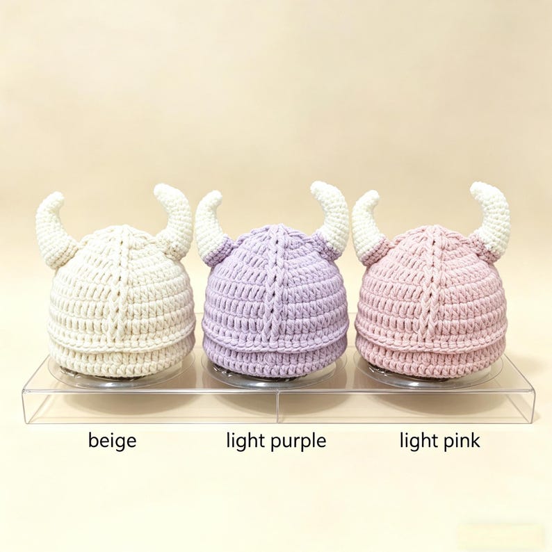 May include: Three crocheted hats with horns, arranged in a row. The hats are colored beige, light purple, and light pink, each featuring white horns. The text below the hats identifies the colors as beige, light purple, and light pink.