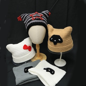 May include: Five crochet cat-ear beanies in various colors and designs. One black and gray striped hat with a red star, a white hat with a red bow, a tan hat with a black patch, and two folded beanies in gray and white with black patches.