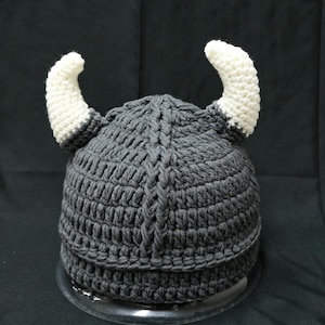 May include: A dark gray crocheted hat featuring a central vertical braided texture and two prominent, curved white horns extending from the sides. The hat is constructed with thick yarn stitches and has a rounded crown with a wide, reinforced brim.
