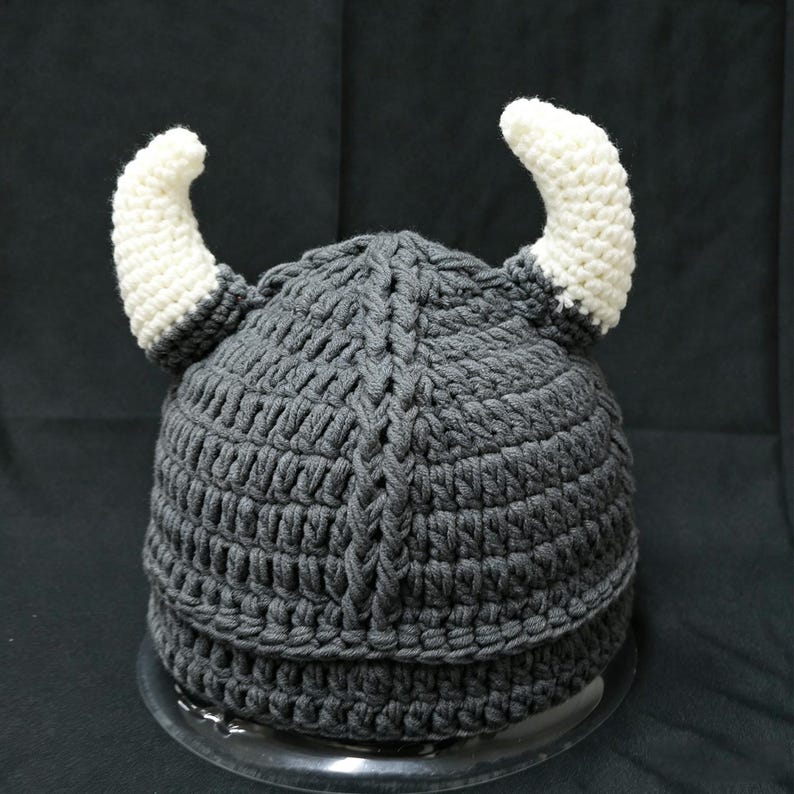 May include: A dark gray crocheted hat featuring a central vertical braided texture and two prominent, curved white horns extending from the sides. The hat is constructed with thick yarn stitches and has a rounded crown with a wide, reinforced brim.