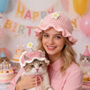 May include: Pink crochet hat with a daisy embellishment, worn by a person and a cat. The cat also wears a matching pink hat. The hats have a ruffled brim and white trim. The background includes birthday decorations.
