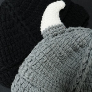 May include: A grey crocheted hat featuring a single white horn protruding from the top. The hat is crafted with a textured yarn stitch pattern and is positioned in front of a solid black crocheted hat, showcasing the contrast in color and design.