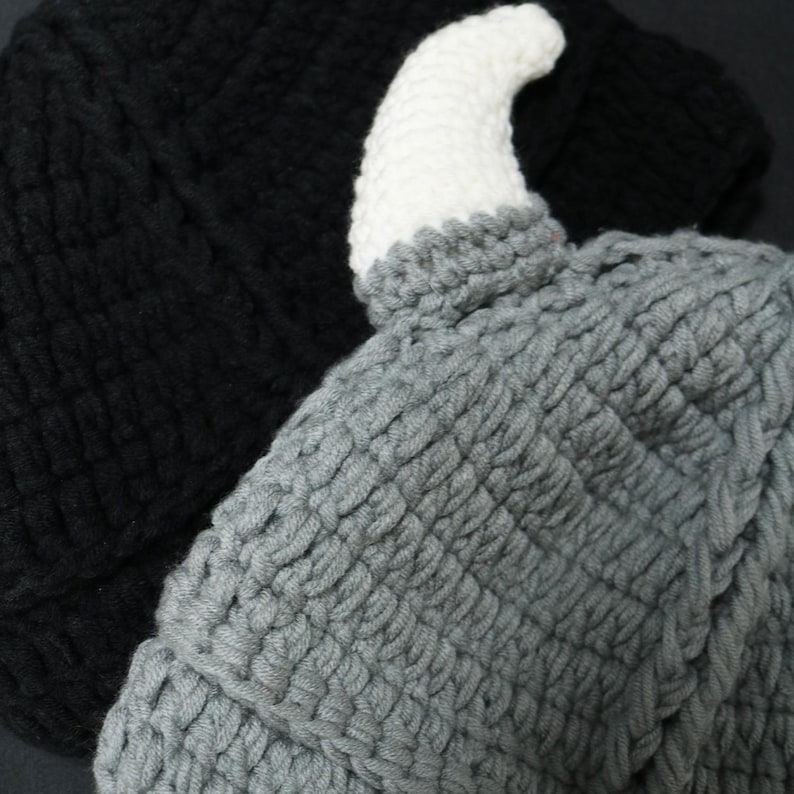 May include: A grey crocheted hat featuring a single white horn protruding from the top. The hat is crafted with a textured yarn stitch pattern and is positioned in front of a solid black crocheted hat, showcasing the contrast in color and design.