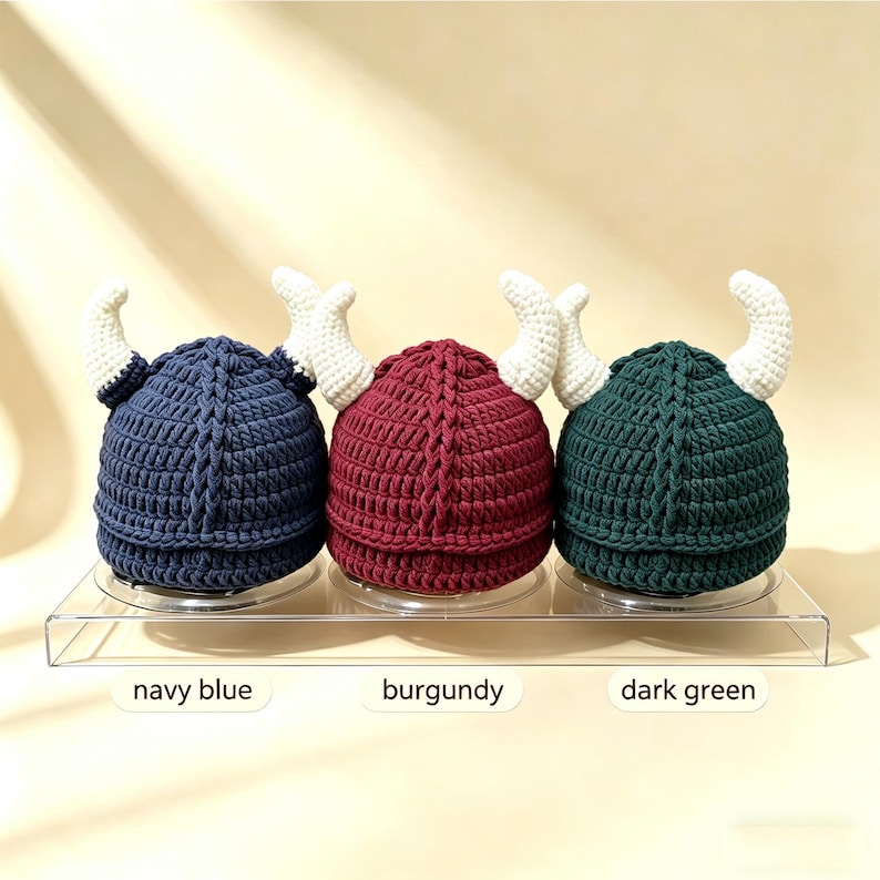 May include: Three crocheted hats in navy blue, burgundy, and dark green, each featuring a central vertical braided texture and two white curved horns extending from the sides. The hats are displayed on a clear stand with labels for each color.