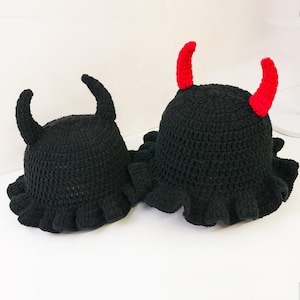 May include: Two black crochet hats with ruffled brims and devil horns. One hat has black horns, and the other has bright red horns. The hats are handmade and sit on a white surface.