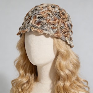 May include: A crochet beanie hat featuring an intricate openwork floral pattern in variegated shades of tan, brown, grey, and cream. The hat has a scalloped edge and is displayed on a mannequin head with long, wavy, light blonde hair.