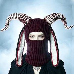 May include: A dark maroon knitted balaclava featuring two large, curved, striped horns in white and maroon. The headgear includes long, dangling ear-like flaps with white centers, maroon borders, and black edges, covering the head and neck area.