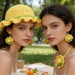May include: Two women wearing matching yellow crochet accessories, including a hat, earrings, and a necklace. The hat has a ruffled brim with green trim. The earrings and necklace feature a sunflower design. The background shows a picnic setting with food and a basket.