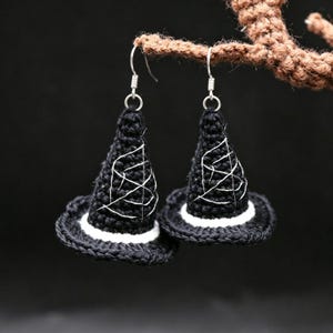May include: A pair of dangling earrings shaped like miniature witch hats. Each hat is crafted from black yarn with a white band at the base and features white embroidered spiderweb detailing on the cone. The earrings hang from silver-toned hooks.