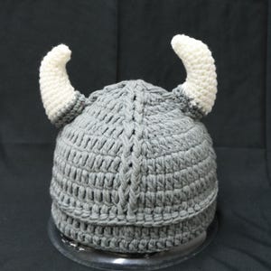 May include: A gray crocheted hat featuring two prominent, curved white horns extending from the sides. The hat is constructed with a thick, textured yarn pattern and includes a vertical braided detail running down the center of the crown.