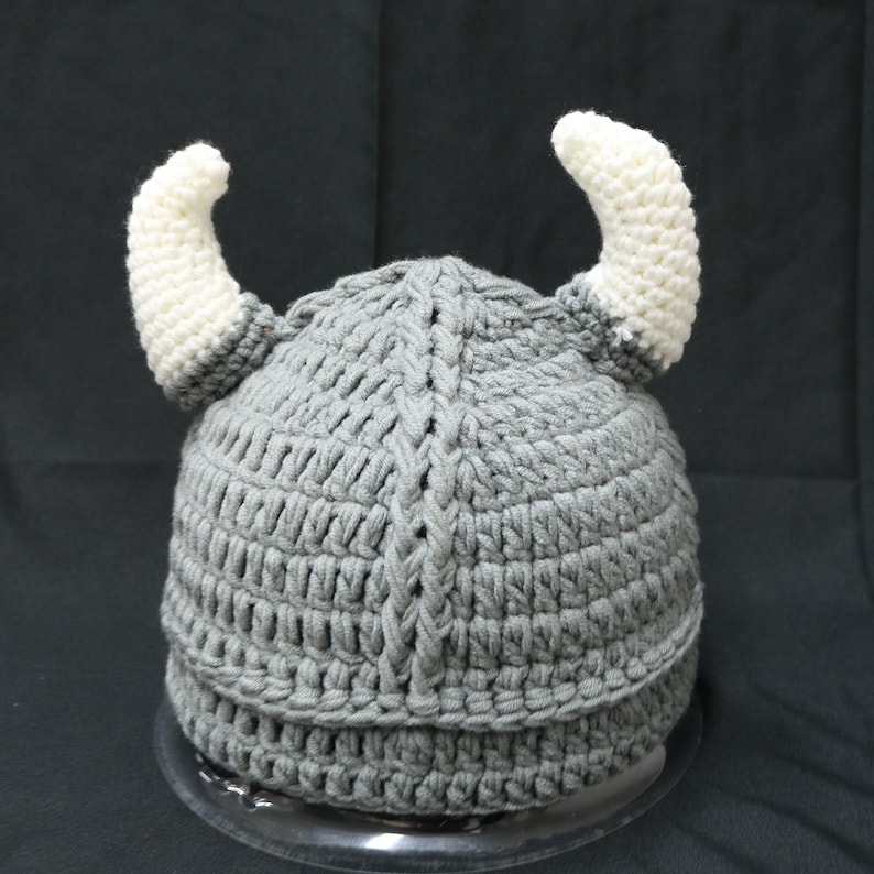 May include: A gray crocheted hat featuring two prominent, curved white horns extending from the sides. The hat is constructed with a thick, textured yarn pattern and includes a vertical braided detail running down the center of the crown.