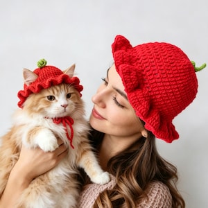 May include: Two matching red crochet hats shaped like strawberries with green stems on top and ruffled brims. One hat is sized for a cat and includes a red chin strap, while the other is a larger version designed for a human to wear.