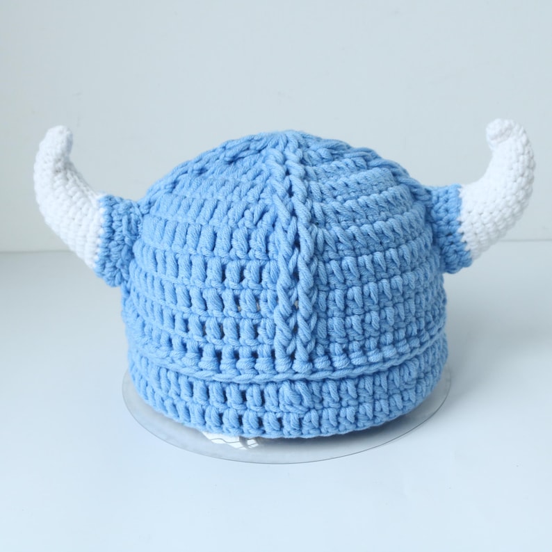May include: A light blue crocheted hat featuring a central vertical ridge and two prominent white crocheted horns extending from the sides. The hat is constructed with a thick yarn texture and has a rounded shape with a wide, ribbed base band.