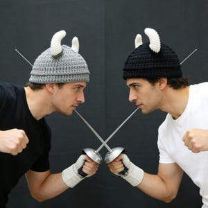 May include: Two crochet hats, one grey and one black, each featuring two white curved horns on top. The hats are constructed with a ribbed texture and are worn by two individuals holding crossed fencing foils with metal guards and white gloves.