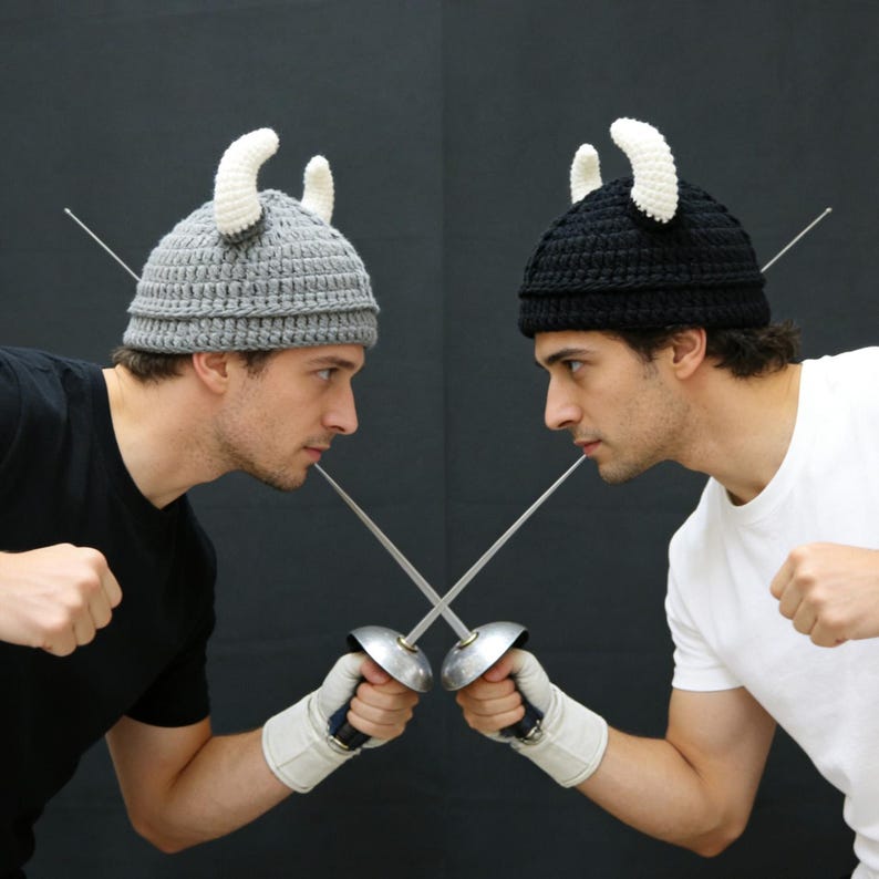 May include: Two crochet hats, one grey and one black, each featuring two white curved horns on top. The hats are constructed with a ribbed texture and are worn by two individuals holding crossed fencing foils with metal guards and white gloves.