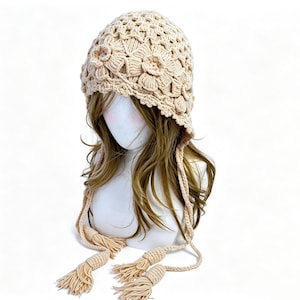 Peut inclure: Un bonnet en crochet beige présentant un motif floral texturé sur tout le tricot. Le bonnet comprend deux longs liens tressés sur les côtés qui pendent vers le bas, chacun terminé par une paire de pompons en fil assortis aux extrémités.