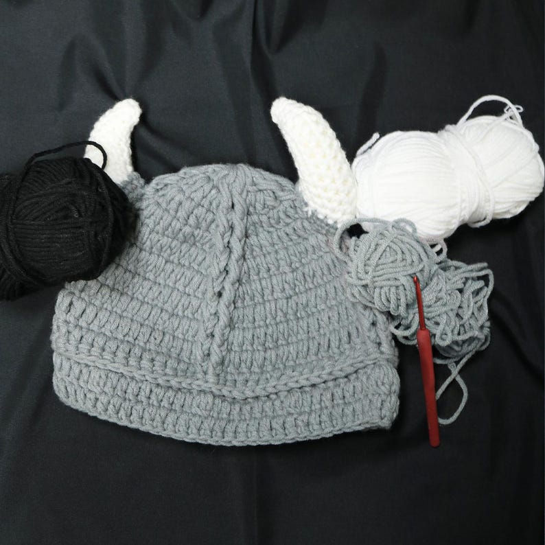 May include: A gray crocheted hat featuring two white horns, accompanied by a ball of black yarn, a ball of white yarn, a loose pile of gray yarn, and a red crochet hook. The hat has a textured, ribbed design with a central vertical braided detail.