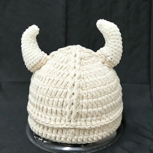 May include: A cream-colored crocheted hat featuring two prominent, curved horns extending from the sides. The hat is constructed with a thick, textured yarn in a vertical ribbed pattern that runs from the crown down to the base edge.