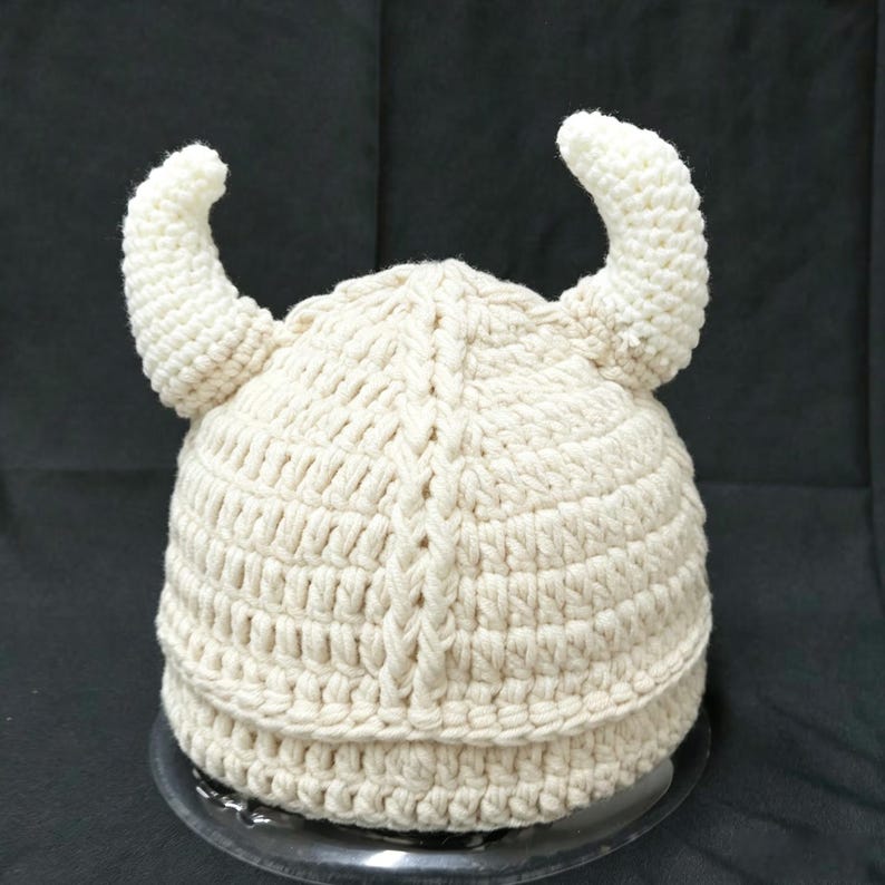 May include: A cream-colored crocheted hat featuring two prominent, curved horns extending from the sides. The hat is constructed with a thick, textured yarn in a vertical ribbed pattern that runs from the crown down to the base edge.