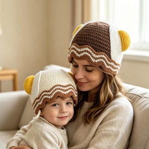 Könnte beinhalten: Zwei passende Häkelmützen mit abgerundeter Krone und kleinen gelben Ohren an den Seiten. Die größere Mütze ist braun mit weißen Akzenten, die kleinere ist weiß mit braunen Akzenten. Beide haben einen gewellten, mehrfarbig gestreiften Rand.