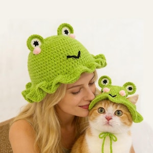 May include: A light green crochet frog hat with a ruffled brim and frog eyes. A cat is wearing a matching hat with a green bow. The cat is being held by a person wearing a matching hat.