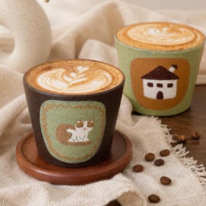 May include: Two ceramic cups filled with latte art. The front cup is dark brown with a light green patch featuring a white and brown cat. The back cup is light green with a tan patch featuring a white house with a brown roof. Both sit on a wooden coaster.