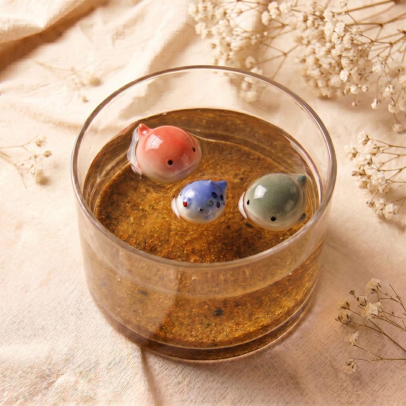 May include: A clear glass bowl filled with amber-colored liquid containing three small, smooth, rounded ceramic animal figurines: one orange, one blue with dark spots, and one pale green, all featuring simple black dot eyes and mouths.