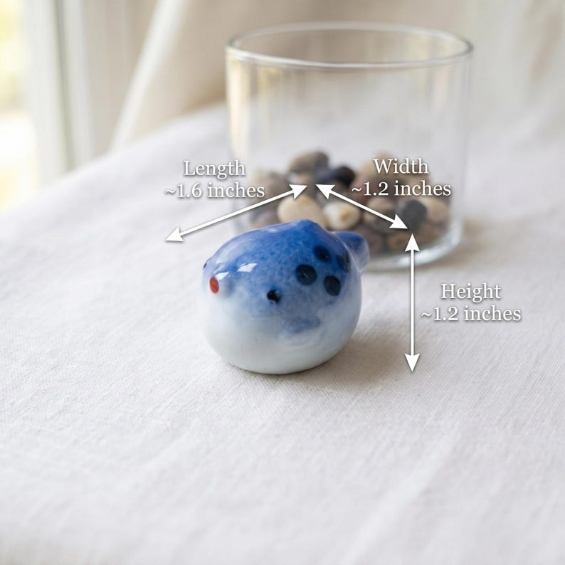 May include: A small, rounded ceramic pufferfish figurine with a blue and white gradient glaze, featuring dark blue spots and a red eye. The figurine measures approximately 1.6 inches in length, 1.2 inches in width, and 1.2 inches in height.