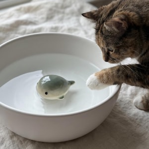 May include: A small, smooth, ceramic whale figurine with a glossy grey and white finish and black eyes, floating in a large, round, white ceramic bowl filled with clear water. The whale has a rounded body, small fins, and a tail.
